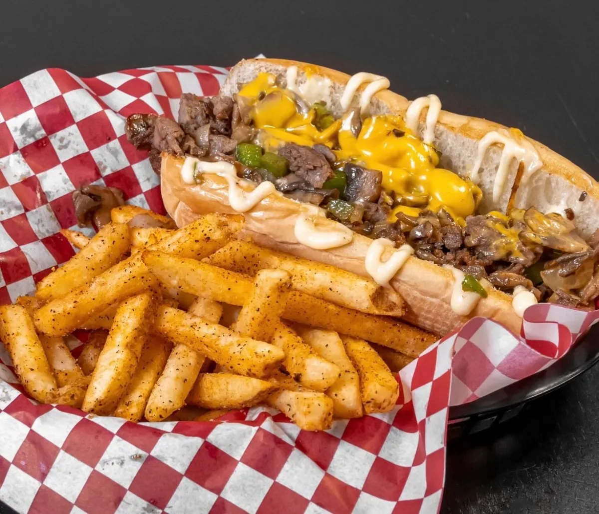 Scorers cheesesteak with peppers and drizzle served with seasoned fries in a red-and-white checkered basket